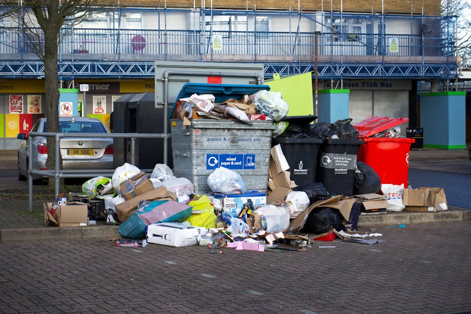 A large pile of waste and rubbish including various black and brown trash bags, cardboard boxes, and loose paper and packaging material, is overflowing from a grey mixed waste bin situated on a paved pavement. The bin, labeled for mixed paper and cardboard, is part of a collection of multiple waste receptacles, including a black bin and a bright red bin, positioned on a street corner. Behind the waste area, there is a metal railing, a silver car parked nearby, and in the background, a building with a blue construction scaffold and signage for a fish bar and other shops. The scene is set during daylight with natural light illuminating the street environment, consistent with private on-site or independent rubbish removal services, such as those provided by Waste Collection Putney, handling non-residential waste disposal in the area.