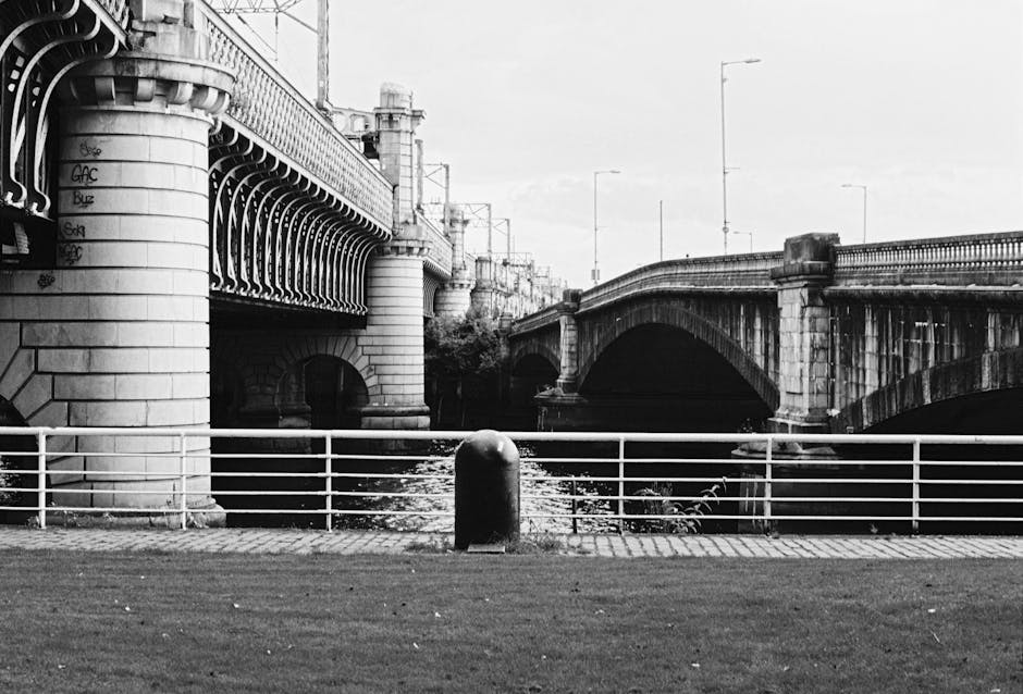A black-and-white photograph of a riverside scene featuring a pedestrian walkway with a metal railing in the foreground. Behind the railing, a short, rounded black bollard is positioned on the paved surface. Directly beyond the railing, the river flows calmly, partially visible beneath two stone bridges with arched spans crossing from left to right. The bridge on the left has a decorative, ornate structure with scrollwork and ironwork details on its side, supported by large stone columns with rounded bases. The bridge on the right appears to be a simpler stone structure with a parapet wall fitted with a row of evenly spaced streetlights on tall poles. In the background, urban buildings and infrastructure are faintly visible, blending into a cloudy sky. The overall scene emphasizes architectural and structural elements typical of urban riverside areas with a focus on classical bridge design and riverside paraphernalia, aligning with a context of private waste management or alternative rubbish collection in the vicinity of the Putney area. The lighting is uniform, with no harsh shadows, highlighting the textures of the stone bridges and the smooth surface of the water.