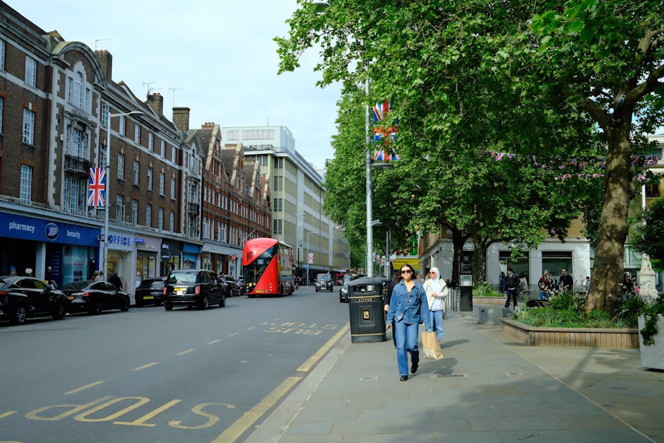 A city street scene with a row of historic brick and stone commercial buildings on the left, featuring retail storefronts including a pharmacy, beauty shop, and office. The storefronts have large display windows, and some have Union Jack flags hanging outside. Several parked cars line the curb, and a red double-decker bus is visible in the middle distance, traveling along the street. On the right side, a wide sidewalk is shaded by mature green-leafed trees, with a few pedestrians walking, including two women dressed in blue academic gowns, one wearing sunglasses, carrying shopping bags. There is a large black trash bin on the sidewalk near a lamppost decorated with Union Jack banners. The environment appears bright and clear, suggesting a daytime scene, with a mixture of older architectural structures and modern buildings further back, all contributing to an active urban atmosphere associated with typical rubbish removal and waste management needs in busy commercial districts.