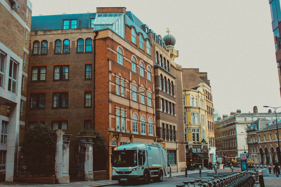 A small street scene showing a white rubbish collection truck parked near the curb in front of a row of historic multi-storey buildings with varied architectural styles. The buildings feature brick facades, large sash windows, and decorative elements, with one displaying an copper-green domed turret. The vehicle is situated on a paved roadway that runs between the buildings, with metal bike racks and bollards arranged along the sidewalk. The environment appears urban with a mix of old and modern structures, and a cloudy sky overhead provides diffuse, natural lighting. This scene reflects a typical setting where private waste collection services, such as those provided by Waste Collection Putney, might operate to handle rubbish removal, especially in historical or densely built city areas where on-site clearance and alternative waste handling options are common.